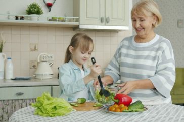 Little girl is cooking salad mixing vegetables in bowl and chatting to cheerful granny in kitchen at home. Modern lifestyle, meals preparation and family concept.