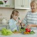 Little girl is cooking salad mixing vegetables in bowl and chatting to cheerful granny in kitchen at home. Modern lifestyle, meals preparation and family concept.